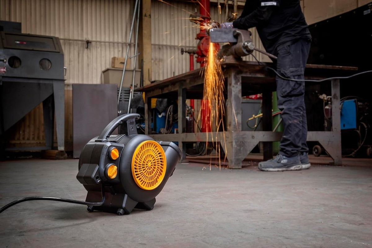 A workshop technician uses a grinding tool on metal, producing sparks near a GEARWRENCH air mover placed on the floor. The blower’s orange front grill is illuminated, indicating active airflow