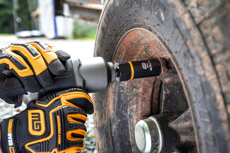 A close-up of a mechanic using an impact gun with a black and orange Bolt Biter extraction socket attached, removing a severely rusted lug nut from a vehicle wheel.