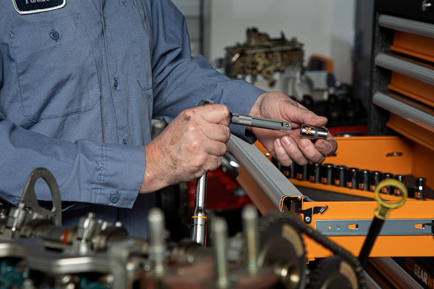 A professional mechanic in a service bay attaching a high-visibility 13mm deep chrome socket onto a 1/2-inch drive extension bar over an open engine compartment.