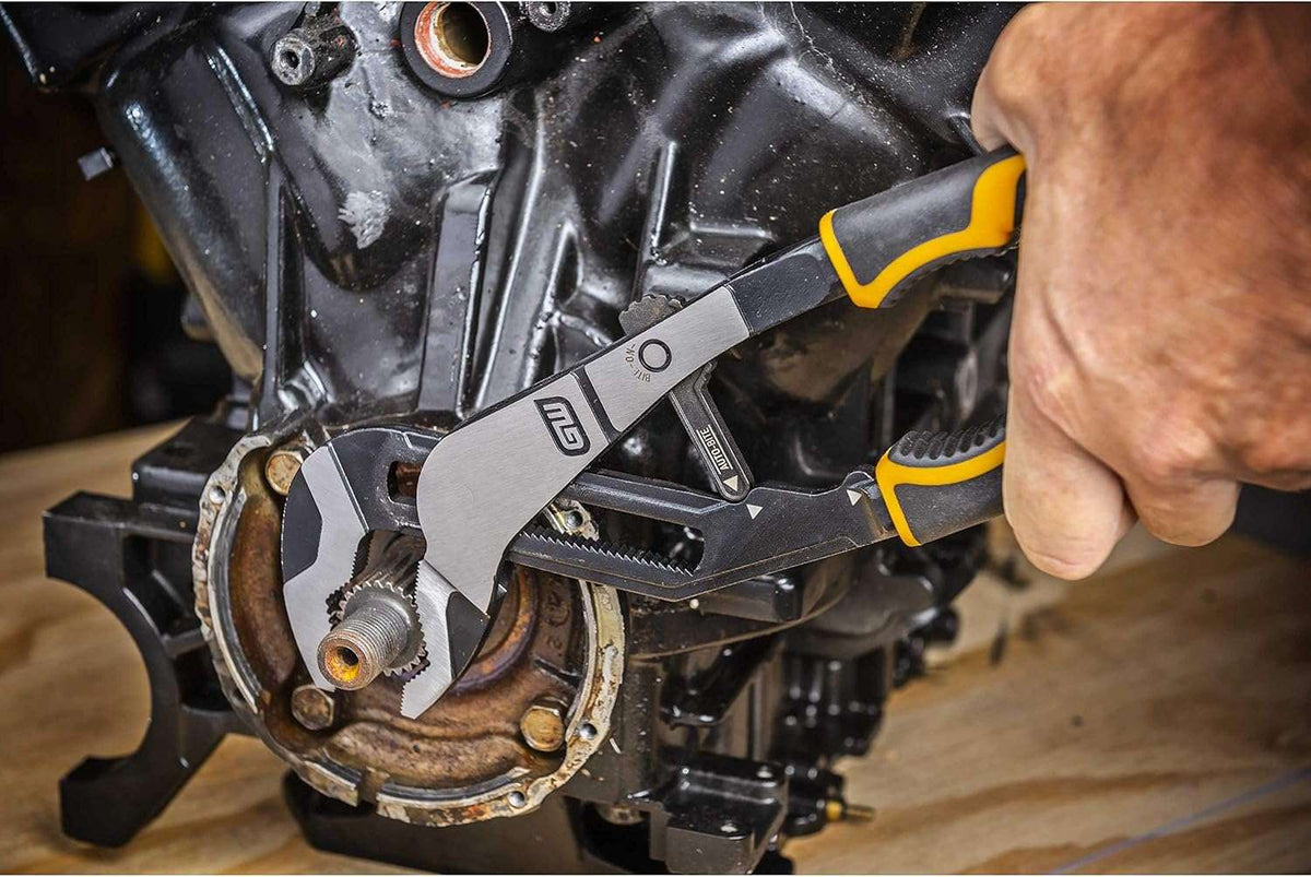 An action shot of the GEARWRENCH PITBULL Auto-Bite pliers being used to grip and turn a large industrial pipe fitting, demonstrating the one-handed Auto-Bite adjustment feature in a real-world application.