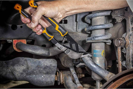 An action shot of the GEARWRENCH 12-inch PITBULL Auto-Bite pliers being used to tighten a large bolt on an industrial machinery component, showcasing the high-leverage grip and one-handed adjustment.