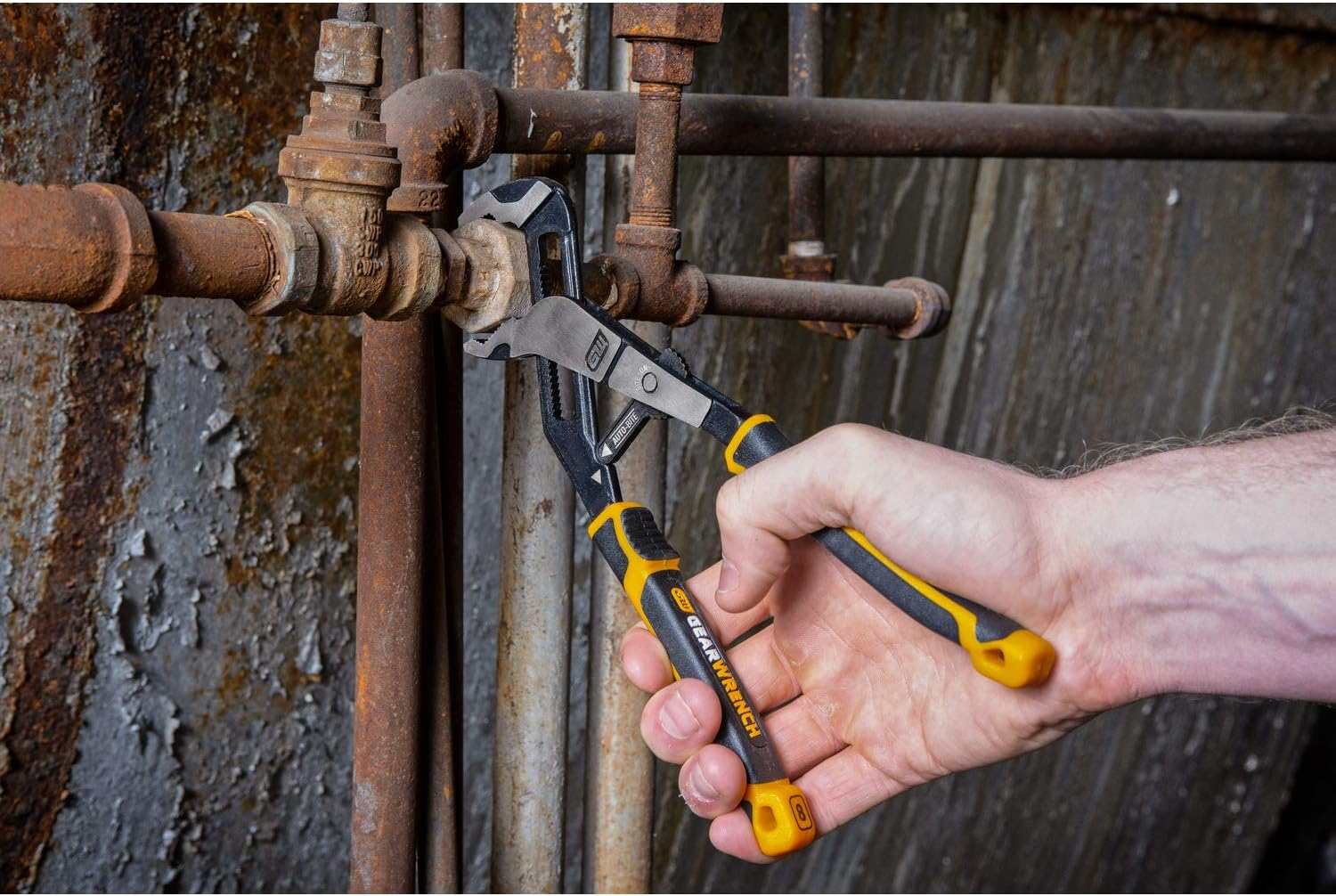 A lifestyle shot of the GEARWRENCH 12-inch Auto-Bite pliers resting on a professional workbench next to industrial fittings, highlighting its rugged alloy steel construction and ergonomic dual-material handles.