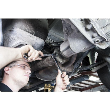 A mechanic wearing safety glasses working underneath a car, using two ratcheting wrenches to loosen a bolt near the exhaust or drivetrain component.