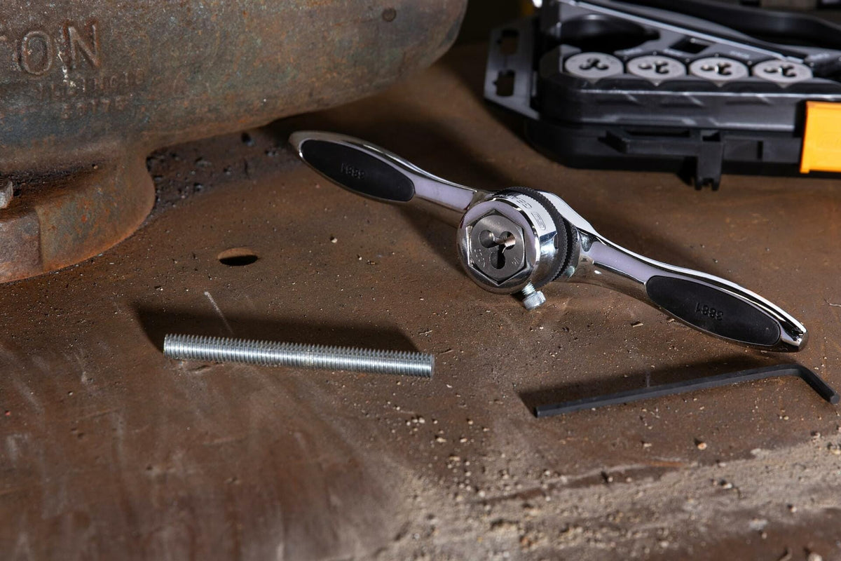 Metalworking setup featuring a die stock handle with black grips labeled “KSEIBI” and a round threading die inserted for cutting external threads. A threaded rod, hex key, and black plastic case with multiple labeled dies are visible on a scratched metal workbench.