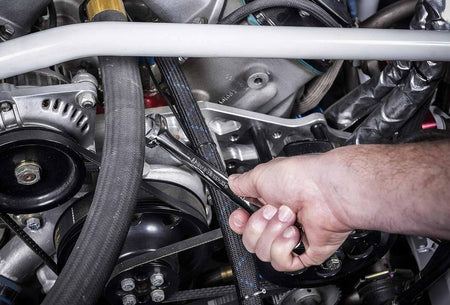 An action photo of a mechanic's hand using a GEARWRENCH long pattern combination SAE wrench to tighten a bolt within a complex vehicle engine bay, showcasing the high-leverage reach and full-polish chrome finish in a professional automotive repair setting.