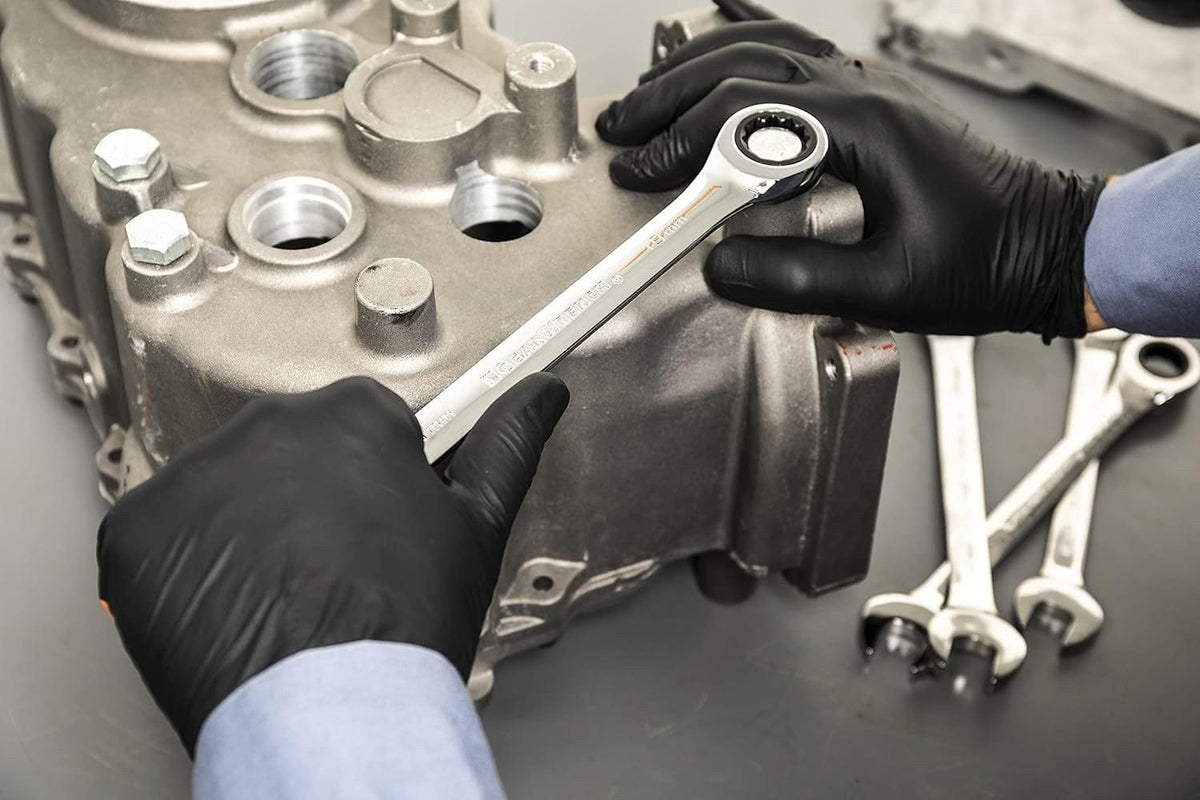 A close-up of a mechanic wearing black gloves using a chrome ratcheting wrench to work on a fastener on a large aluminum transmission or engine casing.