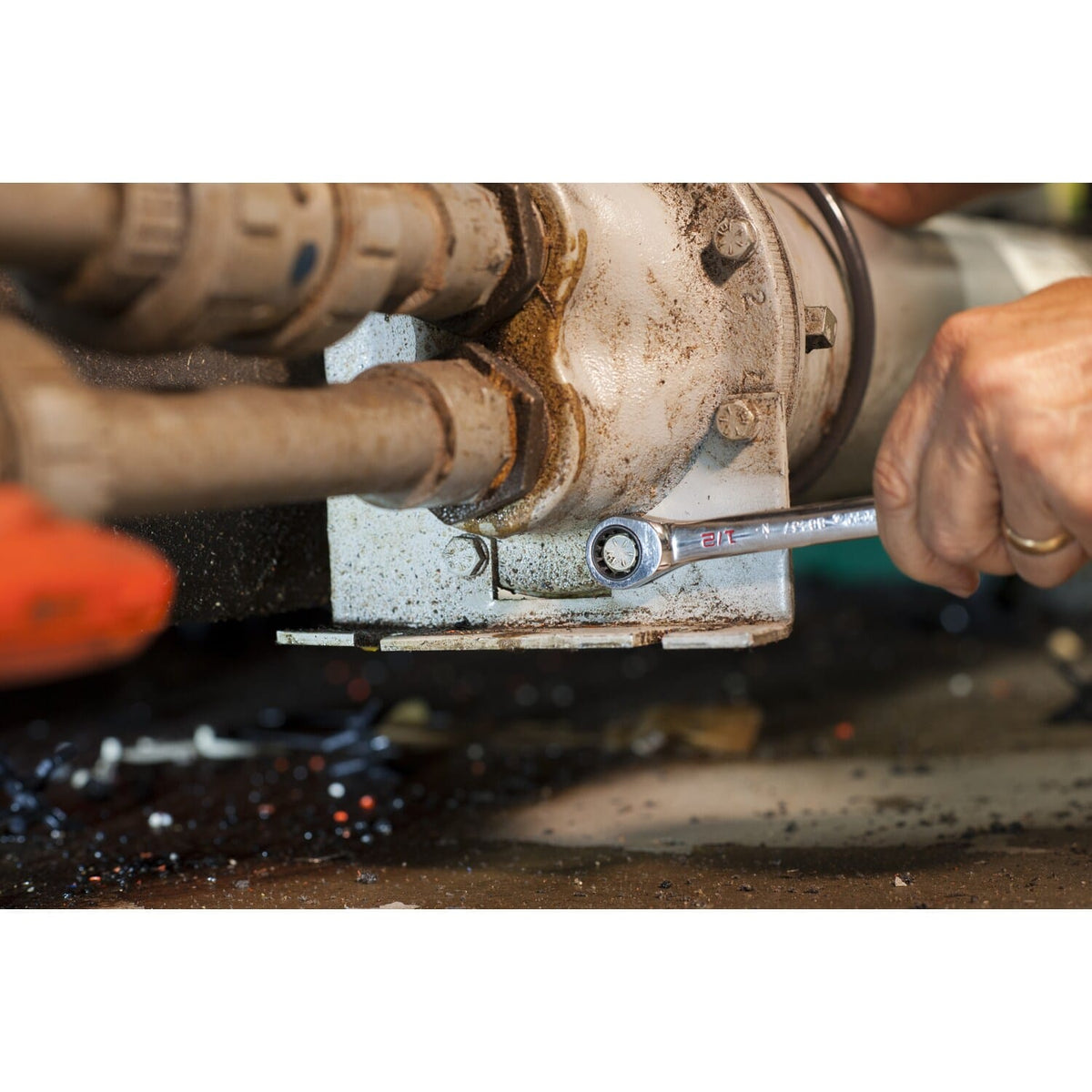 An industrial application shot showing a person using the open end of a GEARWRENCH SAE XL ratcheting combination wrench on a bolt within a complex pipe and flange system.