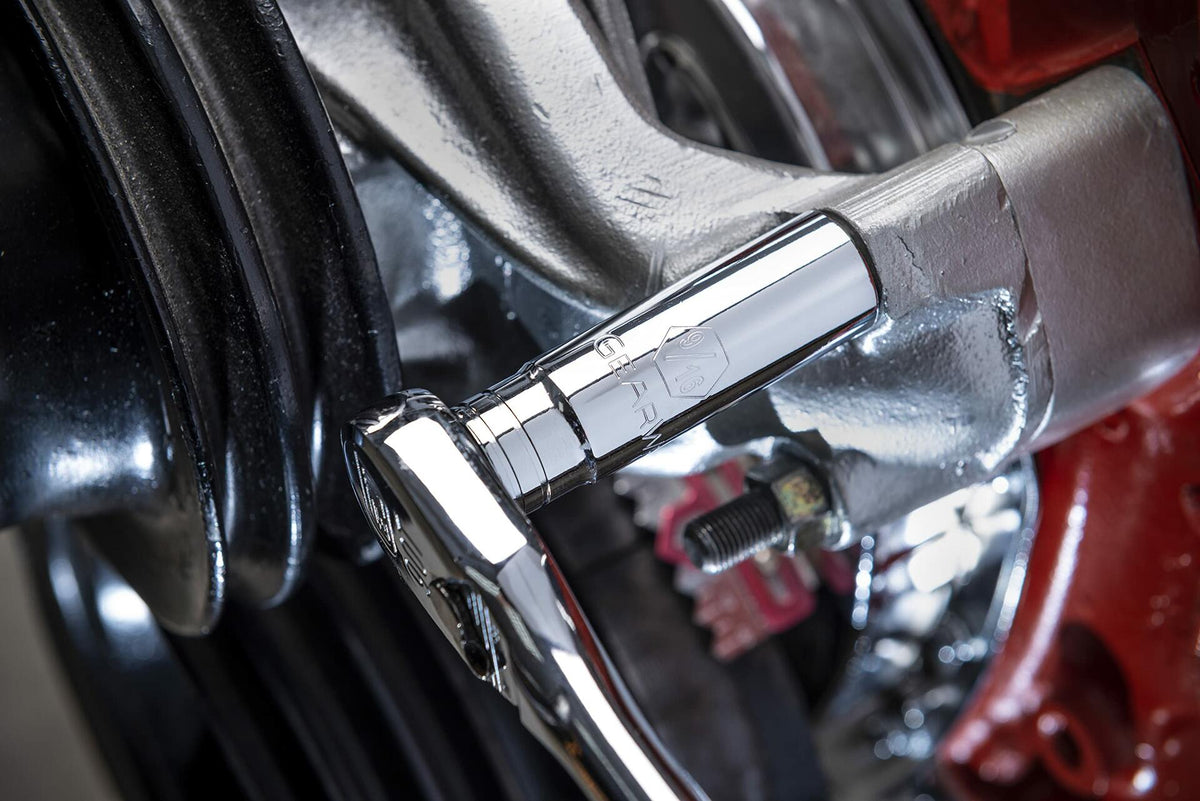 A close-up action shot of a GEARWRENCH 13mm chrome socket and ratchet being used to tighten a bolt on a vehicle's shiny silver brake caliper assembly.