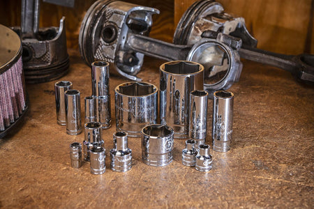 A collection of standard and deep chrome sockets from the GEARWRENCH 89073 set, displayed on a wooden workbench surrounded by engine pistons and an air filter.