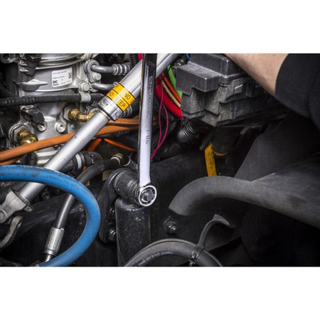 An action photo of a technician's hand using a GEARWRENCH long pattern SAE combination wrench to tighten a bolt on a heavy-duty industrial chassis, showcasing the 12-point box end and full-polish chrome finish in a professional mechanical environment.