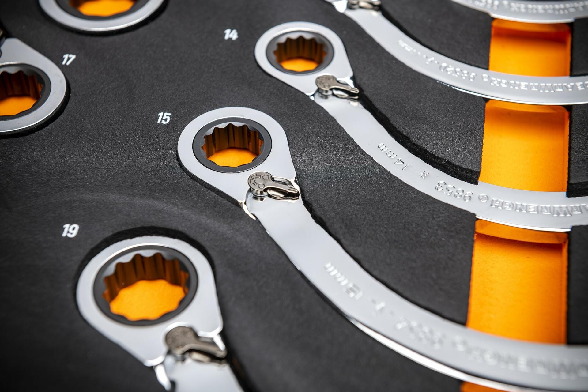 Close-up of ratcheting box-end wrenches in a foam tray labeled with metric sizes including 14, 15, 17, and 19; chrome finish and 12-point ratcheting ends visible against black and orange foam.