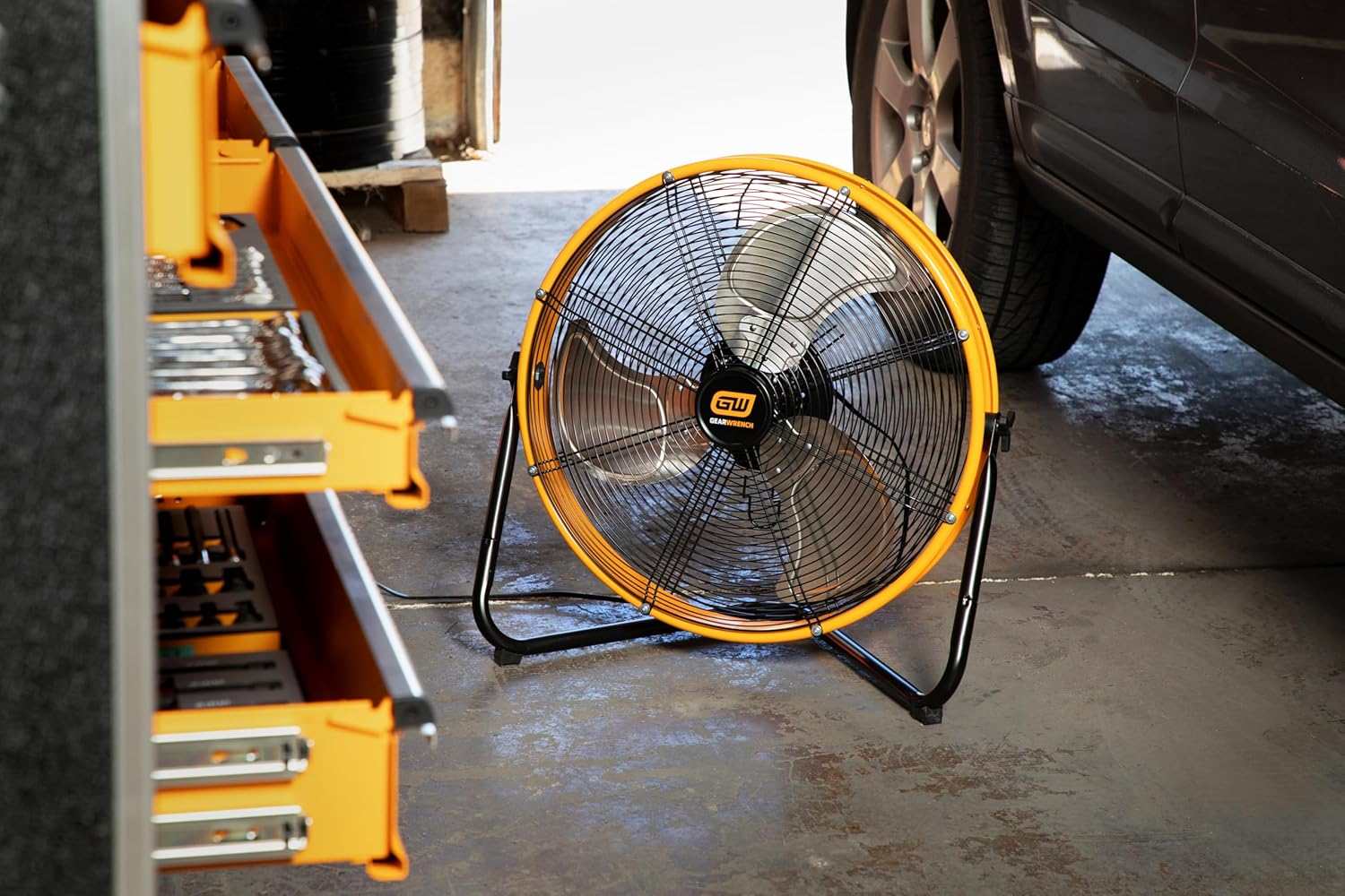 The GEARWRENCH 20-inch 3-speed high velocity industrial shop fan resting on a concrete garage floor next to an open tool chest and a parked vehicle, demonstrating its ideal use in an automotive workspace.