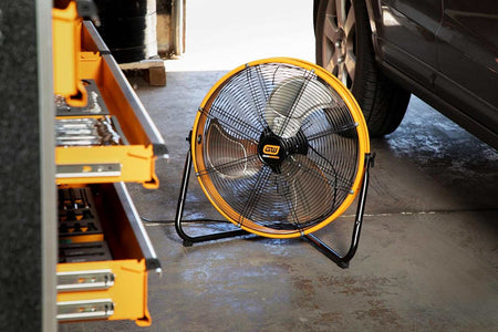 The GEARWRENCH 20-inch 3-speed high velocity industrial shop fan resting on a concrete garage floor next to an open tool chest and a parked vehicle, demonstrating its ideal use in an automotive workspace.