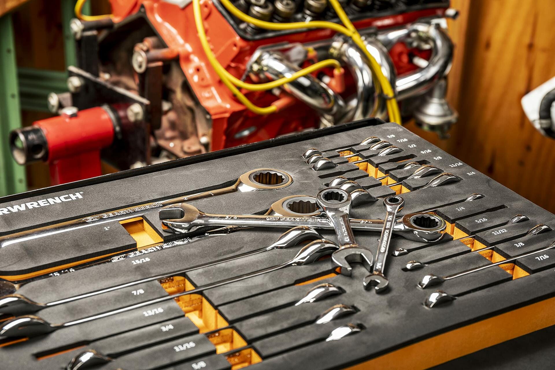 SAE combination wrenches organized in a foam tray labeled with sizes from 1/4 to 7/8 inches. Several wrenches rest on top of the tray. A chrome engine with yellow spark plug wires is visible in the background, suggesting active use in a garage.