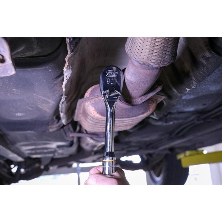 A mechanic's hand holding a chrome 90-tooth ratchet wrench attached to a socket, working underneath a raised vehicle on an exhaust pipe or catalytic converter.