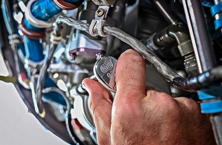 A close-up action shot of a hand using the teardrop head of a GEARWRENCH 90-tooth ratchet to work on a fastener in a complex, tight engine bay surrounded by hoses and fittings.