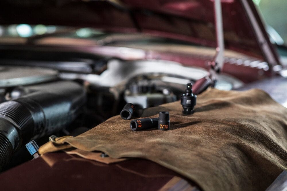 An action photo of GEARWRENCH 3/8 inch drive SAE Hi-Viz impact sockets and a universal joint resting on a protective fender cover of a classic vehicle, illustrating the tool set's use in professional automotive restoration and maintenance.