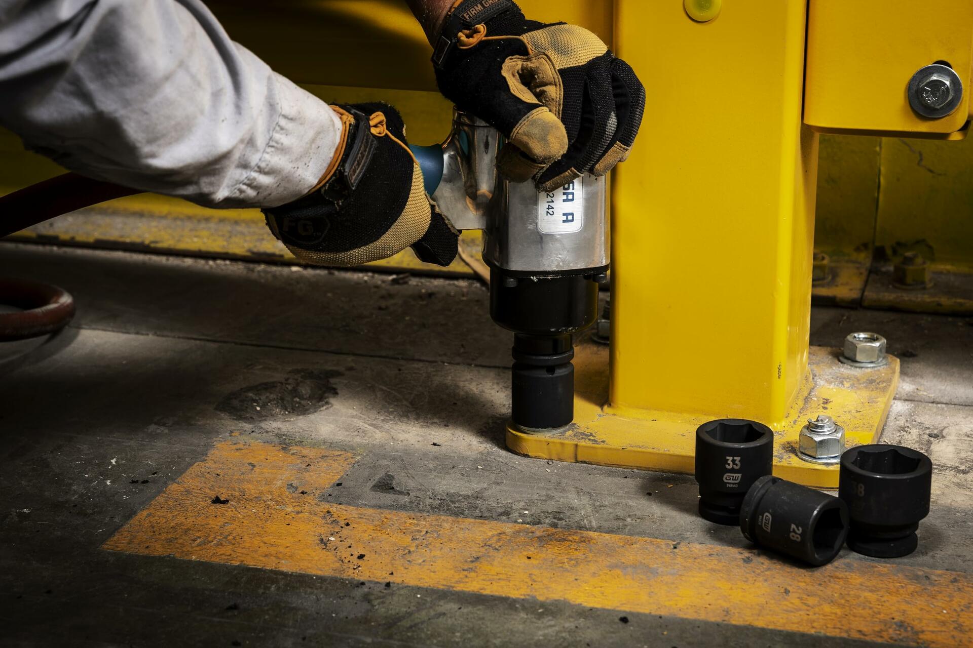 Person using a pneumatic impact wrench to tighten a bolt on a yellow industrial structure. Several black impact sockets labeled with sizes like “33” and “36” are scattered nearby. The scene is set on a concrete floor with yellow safety markings, indicating a professional workshop or construction site.