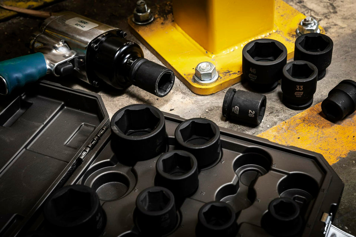 Impact sockets and a pneumatic impact wrench placed on a concrete floor near a yellow industrial structure. Socket sizes include 46mm, 33mm, 29mm, and 28mm. Tools are arranged around a molded plastic case, illustrating real-world use in heavy-duty environments.