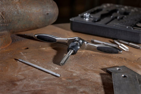 Workshop scene showing an IRWIN T-handle tap wrench holding a threading tap positioned on a metal surface. Surrounding tools include a black case with taps and dies, a screwdriver bit, and a rusted metal object in the background. Demonstrates threading setup in a metalworking environment.