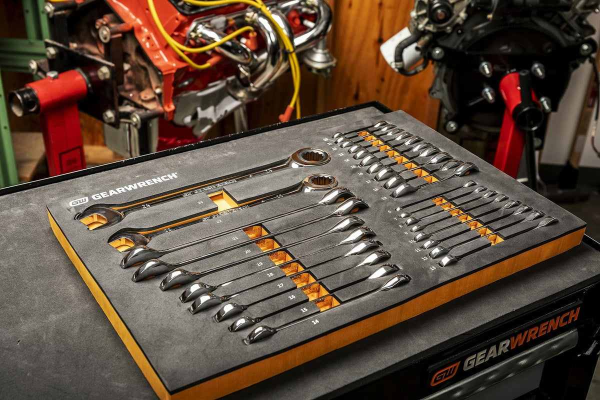 A GEARWRENCH foam tray on a black workbench holds a set of combination wrenches with orange highlights. Two engine blocks on red stands are visible in the background, indicating active use in a workshop.