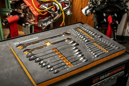 A GEARWRENCH foam tray on a black workbench holds a set of combination wrenches with orange highlights. Two engine blocks on red stands are visible in the background, indicating active use in a workshop.