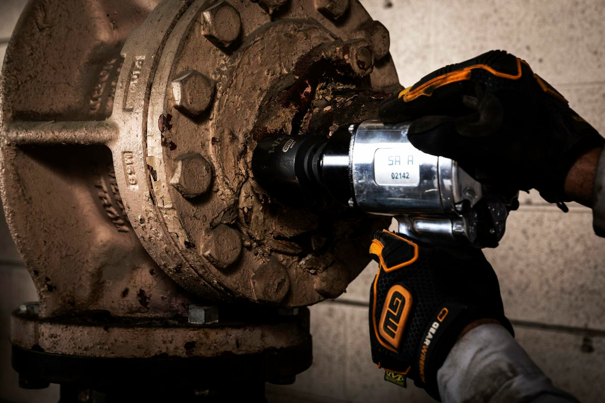 A mechanic wearing GEARWRENCH gloves uses an air impact wrench with a socket to remove a large, rusty bolt from an industrial flange.