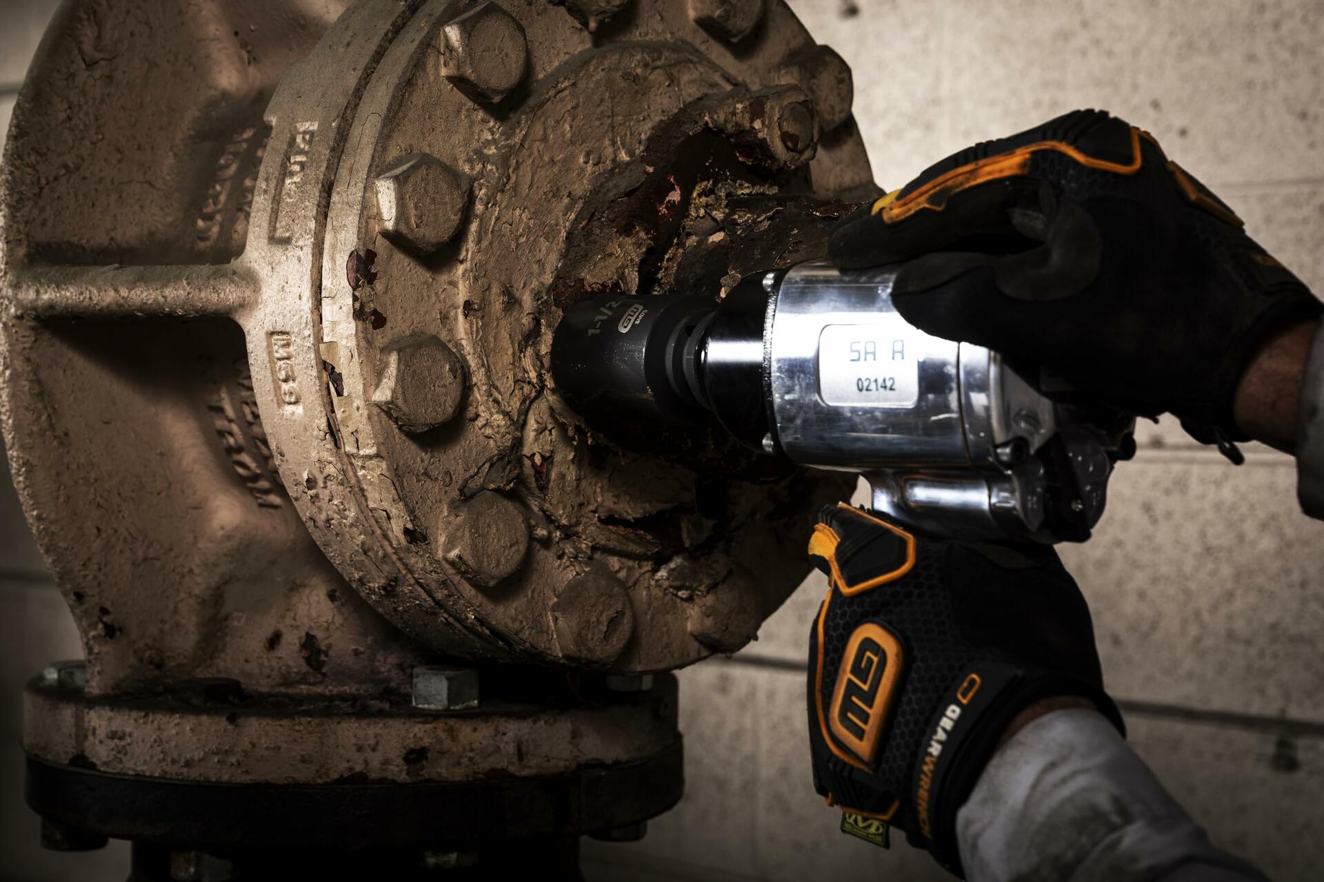 A mechanic wearing GEARWRENCH gloves uses an air impact wrench with a socket to remove a large, rusty bolt from an industrial flange.