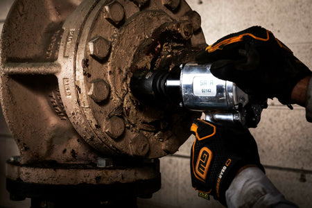 A mechanic wearing GEARWRENCH gloves uses an air impact wrench with a socket to remove a large, rusty bolt from an industrial flange.