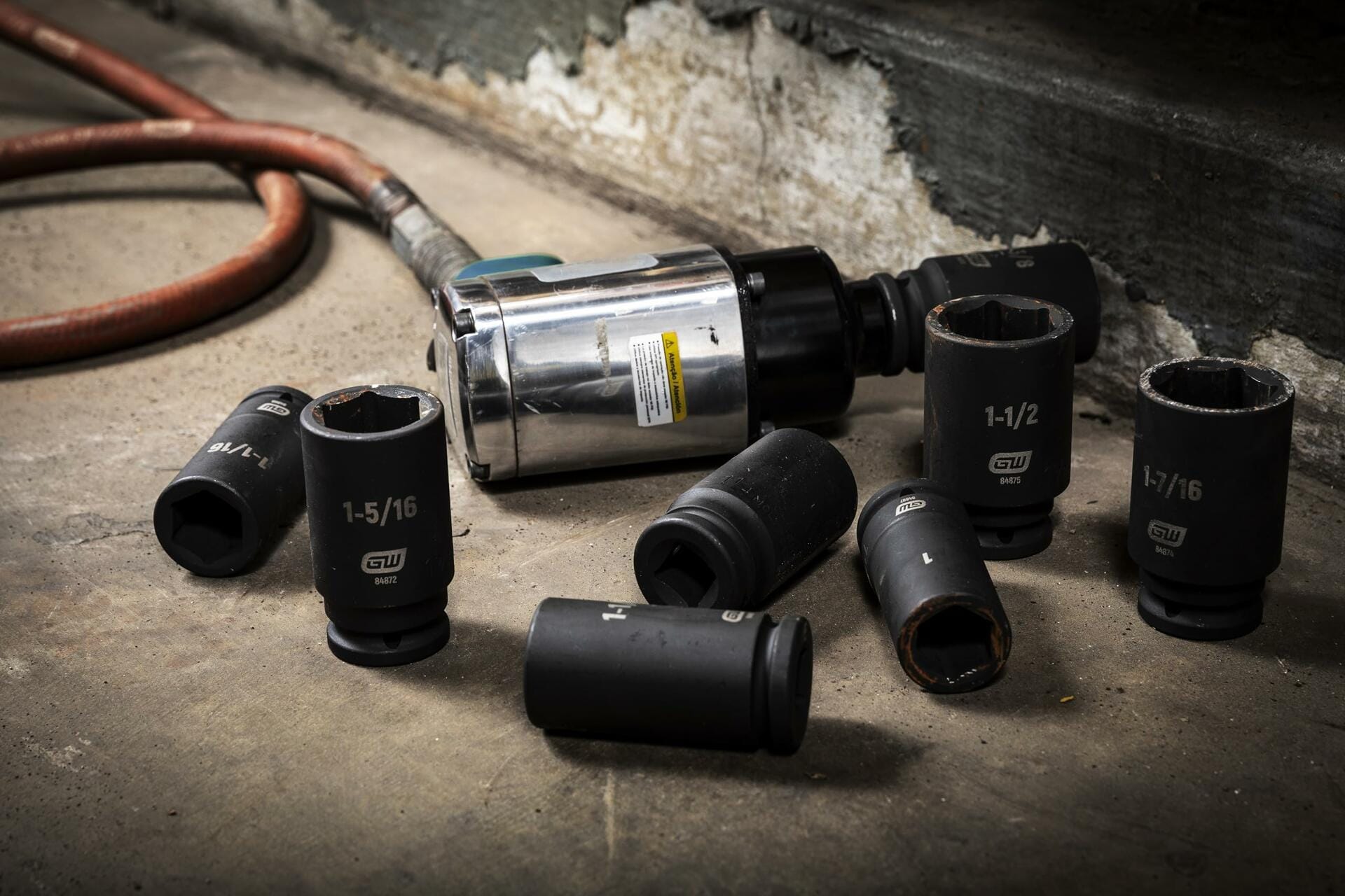 Several black GEARWRENCH deep impact sockets, including 1-5/16 and 1-1/2 inch sizes, lying on a concrete shop floor next to a pneumatic impact wrench and air hose.