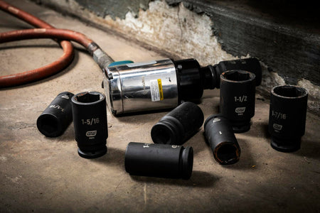 Several black GEARWRENCH deep impact sockets, including 1-5/16 and 1-1/2 inch sizes, lying on a concrete shop floor next to a pneumatic impact wrench and air hose.
