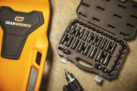 The open GEARWRENCH 84948N impact socket set resting on a shop floor next to an impact wrench and a yellow GEARWRENCH creeper.