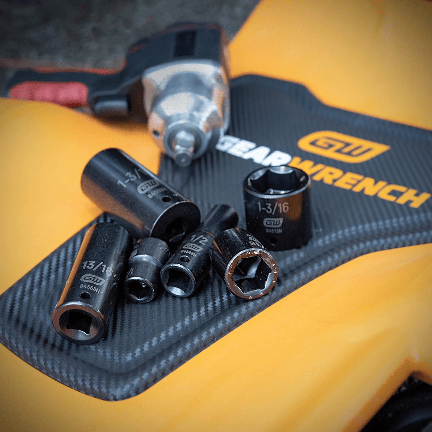 A collection of GEARWRENCH 1/2-inch drive impact sockets, including sizes 13/16, 1-3/16, and 1/2, resting on a yellow and black GEARWRENCH creeper with an impact wrench blurred in the background.