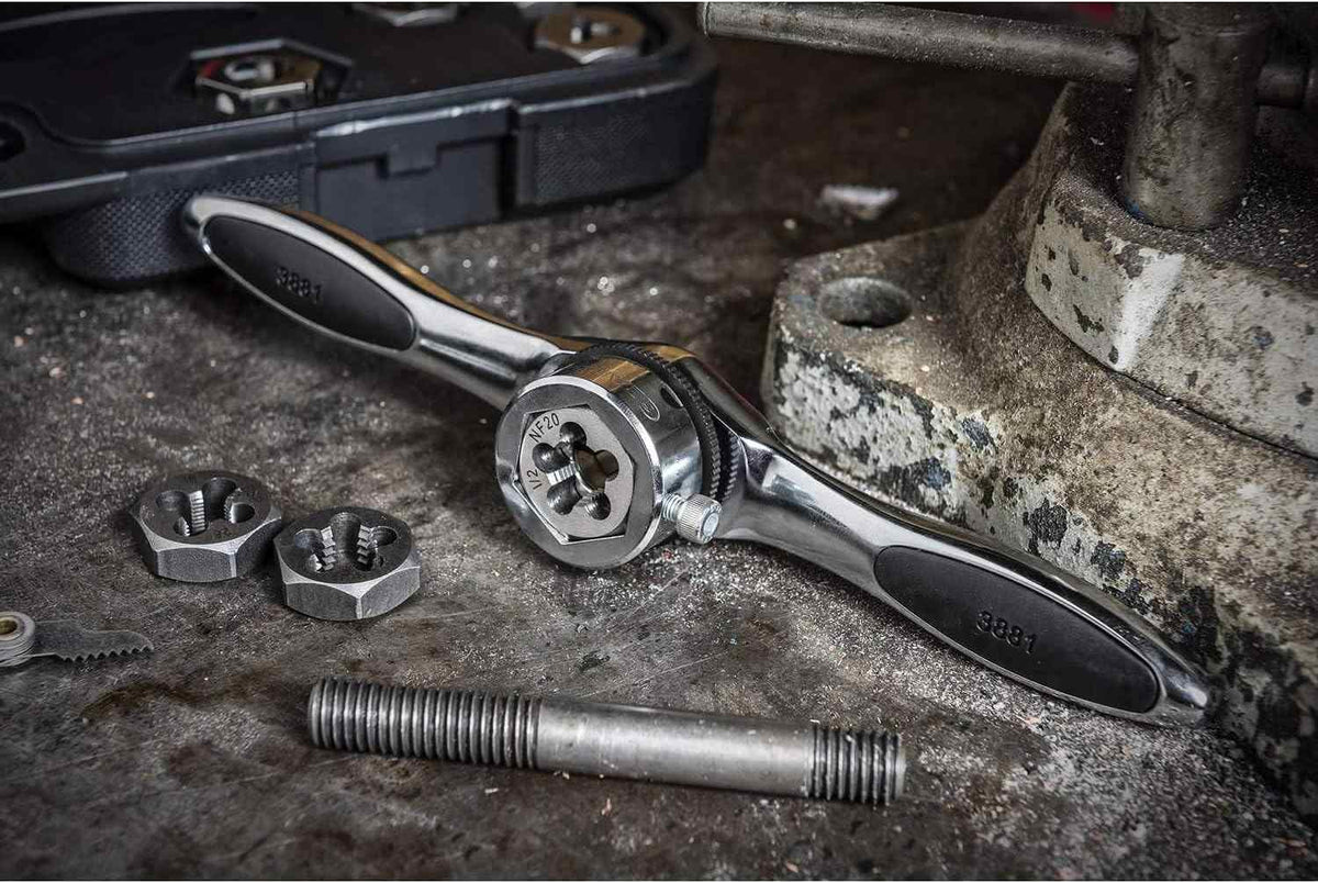 Lifestyle shot of the GearWrench ratcheting T-handle and hex dies on a metal workbench next to a vise, demonstrating its use in cleaning up damaged threads on a large steel bolt.