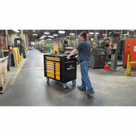 A factory worker rolling the black and orange GEARWRENCH 11-drawer mobile work station down an aisle in an industrial manufacturing facility.