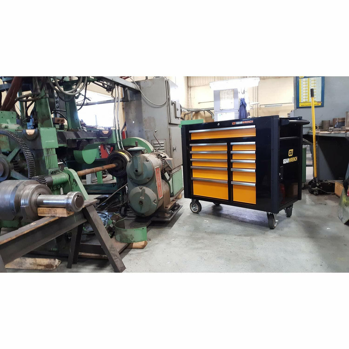 The black and orange GEARWRENCH 11-drawer mobile work station parked on the floor of a machine shop or industrial facility, positioned near large machinery.