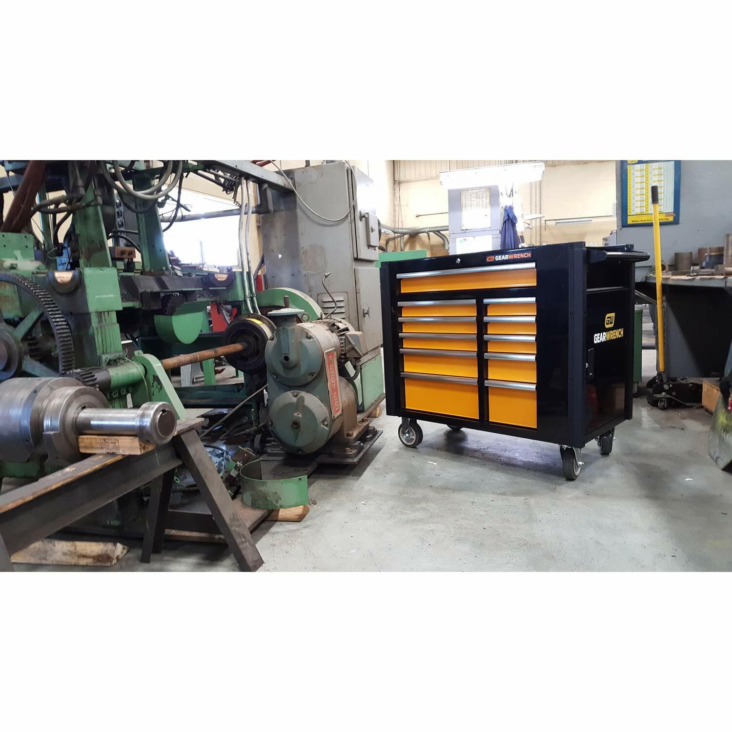 The black and orange GEARWRENCH 11-drawer mobile work station parked on the floor of a machine shop or industrial facility, positioned near large machinery.