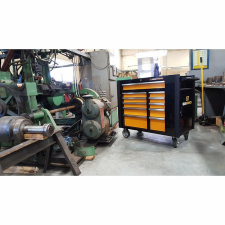 The black and orange GEARWRENCH 11-drawer mobile work station parked on the floor of a machine shop or industrial facility, positioned near large machinery.