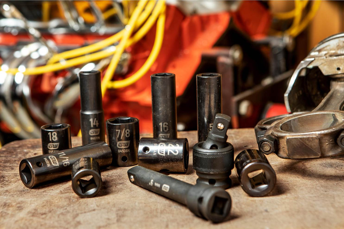 A lifestyle shop shot of various GEARWRENCH SAE and metric impact sockets, an extension bar, and a universal joint scattered on a wooden workbench in front of a vehicle engine, demonstrating real-world professional use.