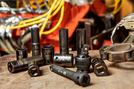 A lifestyle shop shot of various GEARWRENCH SAE and metric impact sockets, an extension bar, and a universal joint scattered on a wooden workbench in front of a vehicle engine, demonstrating real-world professional use.