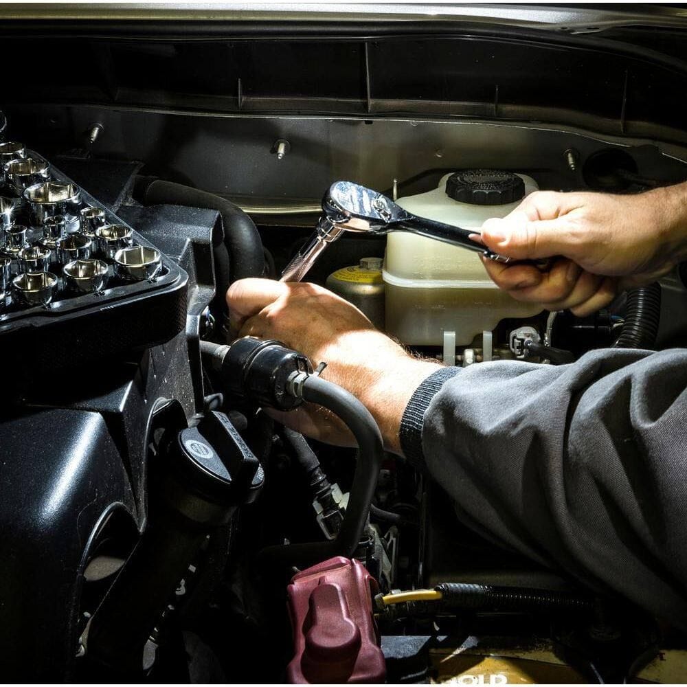 Action shot of a professional mechanic using the GearWrench 90-tooth 1/2-inch drive ratchet and socket set to perform maintenance inside a vehicle engine bay under bright workspace lighting.