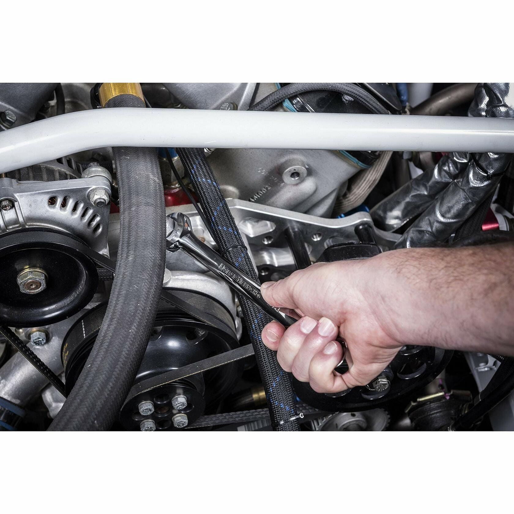 Close-up of a hand using a wrench to adjust a component in a car engine bay. The wrench is applied to the serpentine belt system near pulleys and hoses, illustrating real-world use of a combination wrench in automotive maintenance.