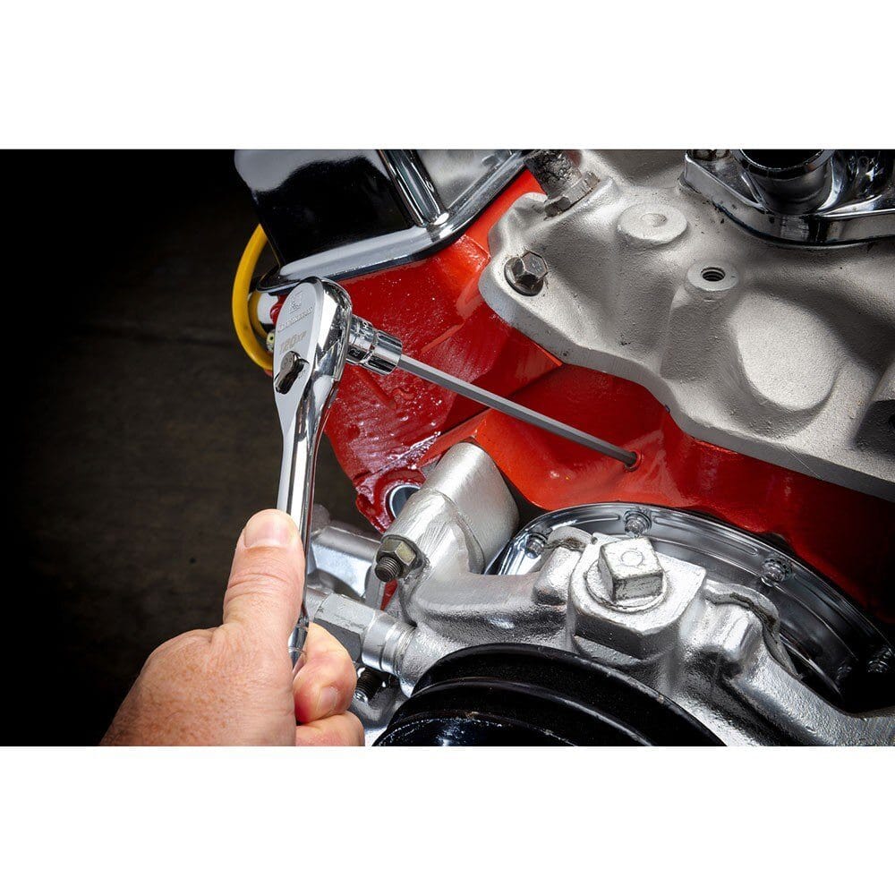 A person's hand using a ratchet wrench attached to a long hex bit socket, reaching a deeply recessed bolt on a red and chrome car engine block.