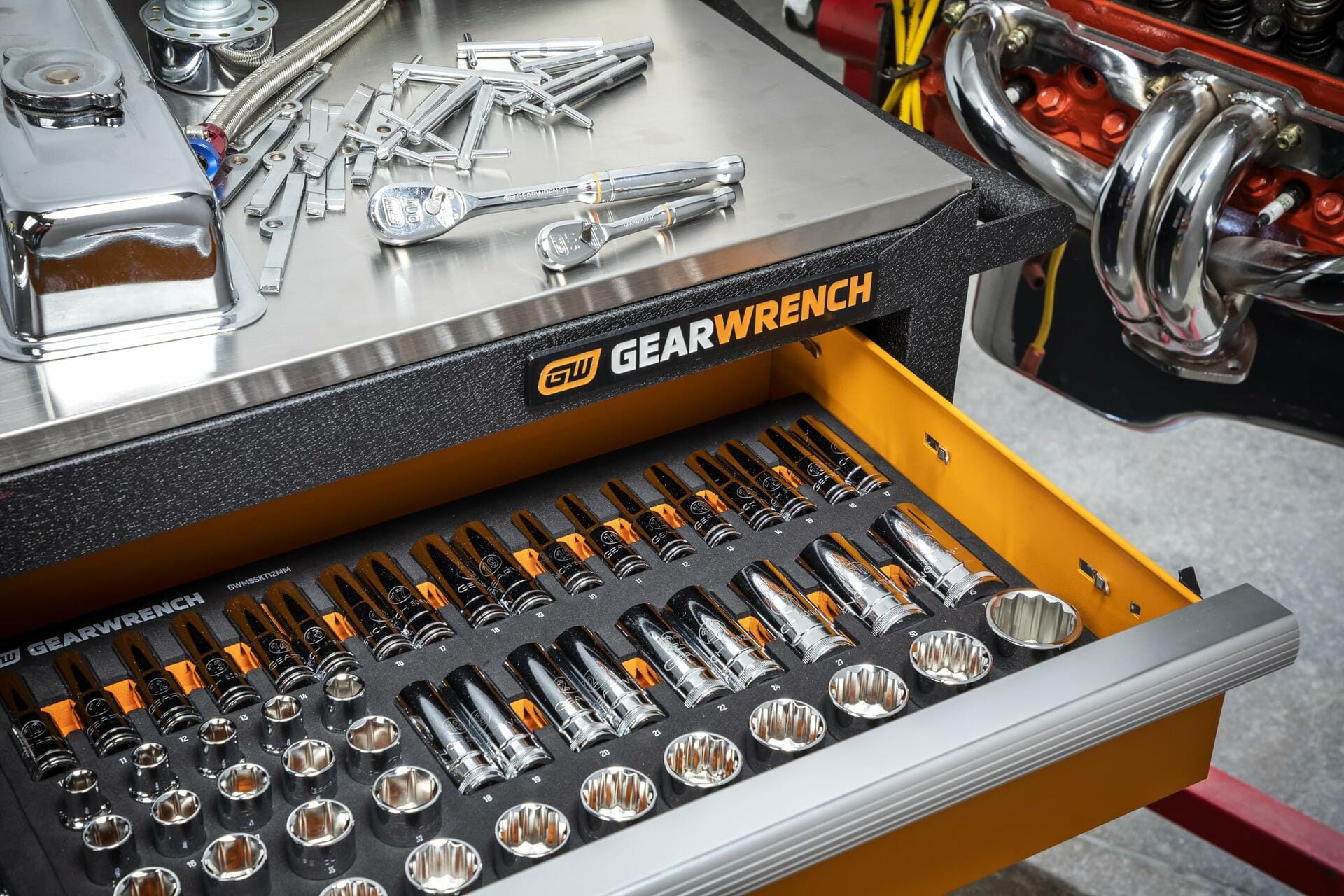 A mechanic’s stainless steel workbench with a GEARWRENCH-branded drawer containing ratchets, socket wrenches, and extensions in a foam insert. Additional tools and a visible engine block suggest active automotive repair use.