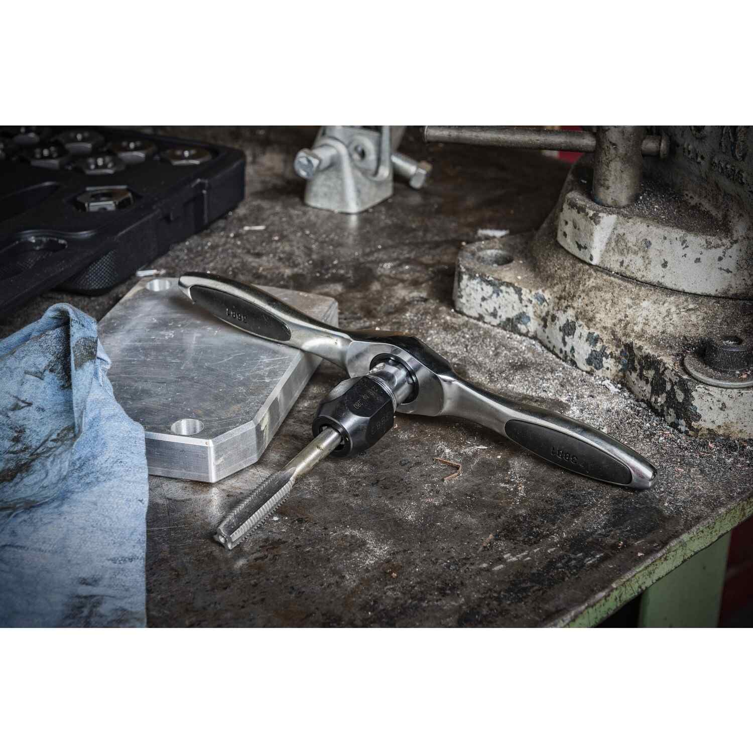 Tap wrench with inserted tap tool on a workshop bench, surrounded by metal components and tools, illustrating threading operations in industrial settings.