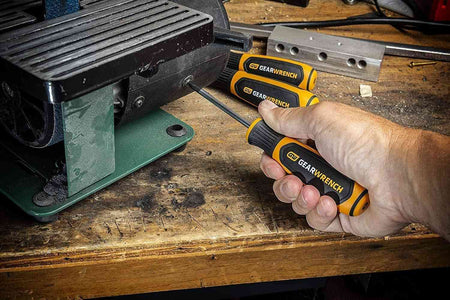 A professional using a GEARWRENCH slotted screwdriver to perform maintenance on a piece of machinery on a wooden workbench, showcasing the tool's precision and comfort in a shop setting.