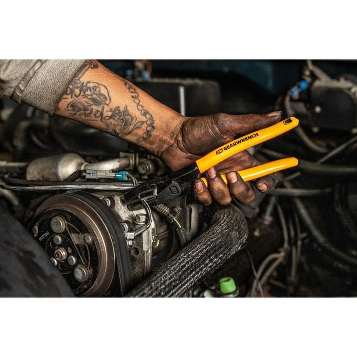 A professional mechanic using the GEARWRENCH PITBULL lineman's pliers to perform maintenance on a vehicle engine, demonstrating the high-leverage grip in a tight workspace.