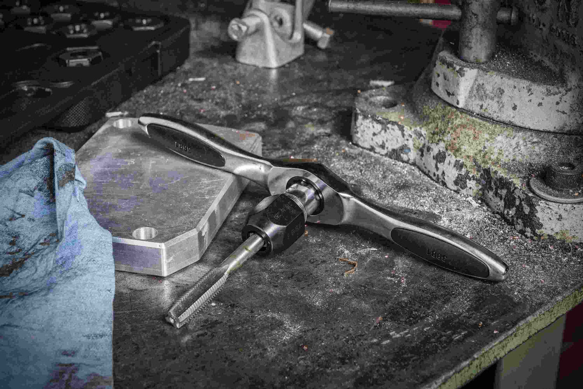 A GEARWRENCH ratcheting T-wrench with a tap adapter and large tap being used on a metal plate, illustrating the Twist Lock guide system that keeps dies and taps centered during use.