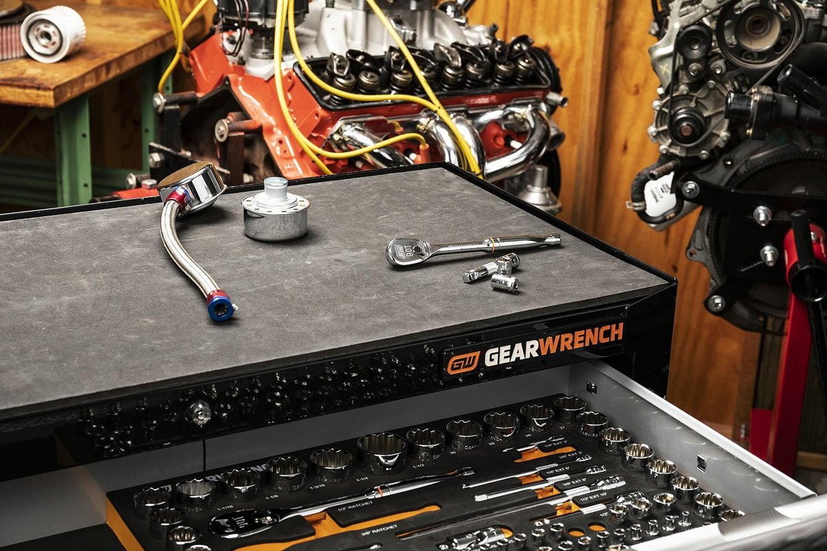 Professional mechanic’s workspace featuring a GEARWRENCH tool chest with open drawer showing organized sockets and hand tools. The top surface holds a ratchet, socket, extension bar, and metal component. Background includes two engine blocks on stands.