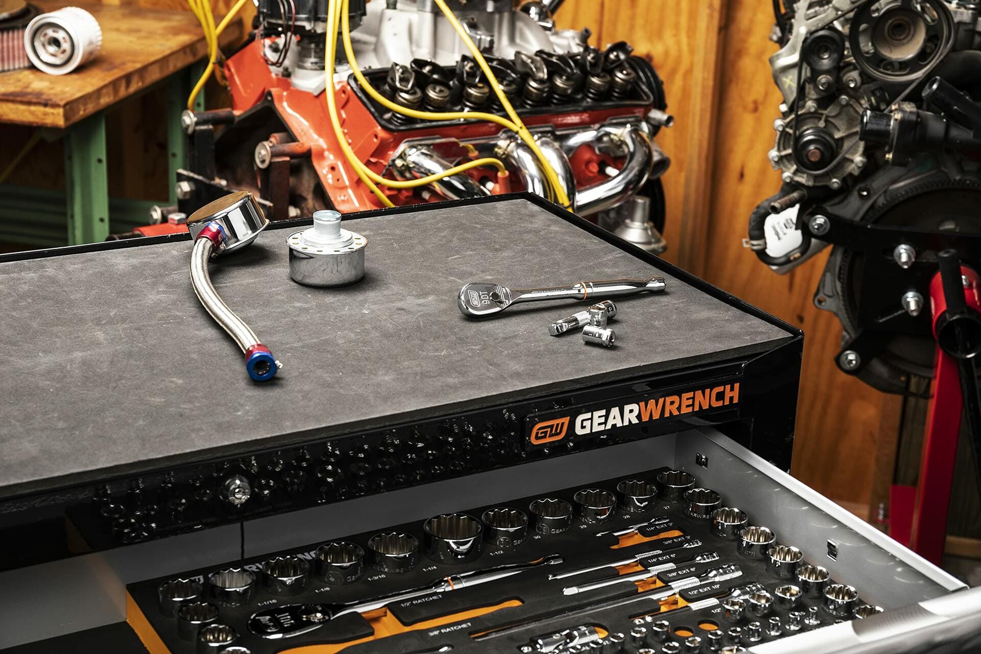 Professional mechanic’s workspace featuring a GEARWRENCH tool chest with open drawer showing organized sockets and hand tools. The top surface holds a ratchet, socket, extension bar, and metal component. Background includes two engine blocks on stands.